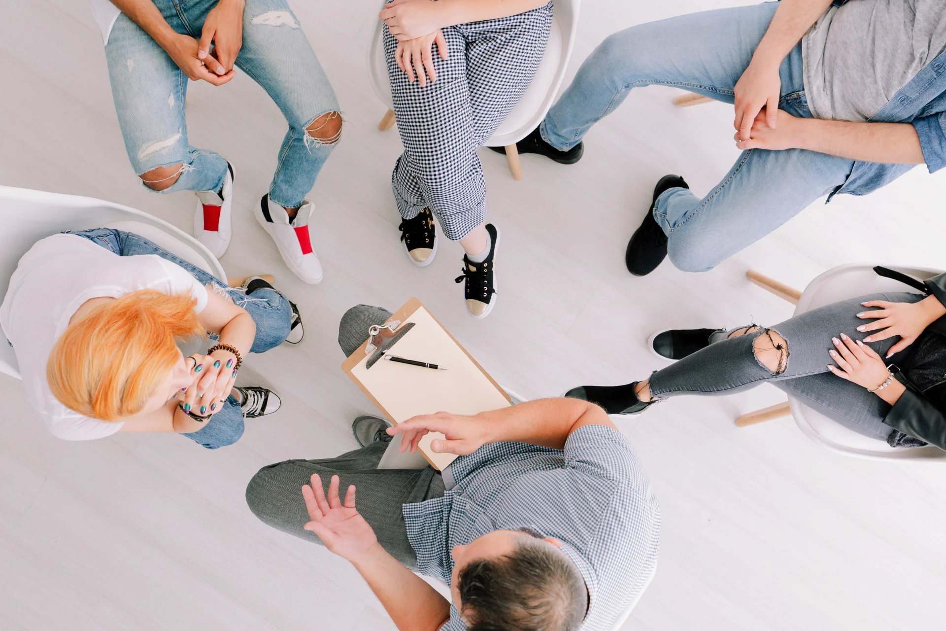 Group therapy session for addiction recovery, participants seated in a circle, engaged in discussion, clipboard with notes visible, promoting support and structured treatment.