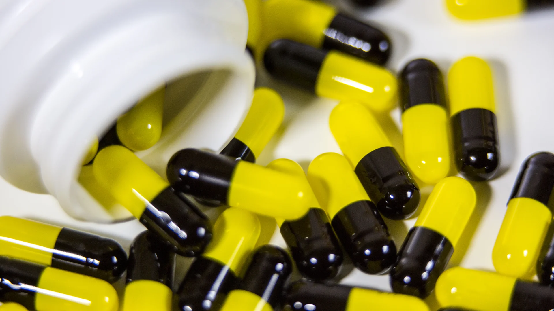 Yellow and black capsules spilled from a white pill bottle, representing medications like Darvocet and Darvon linked to opioid dependence and addiction risks.