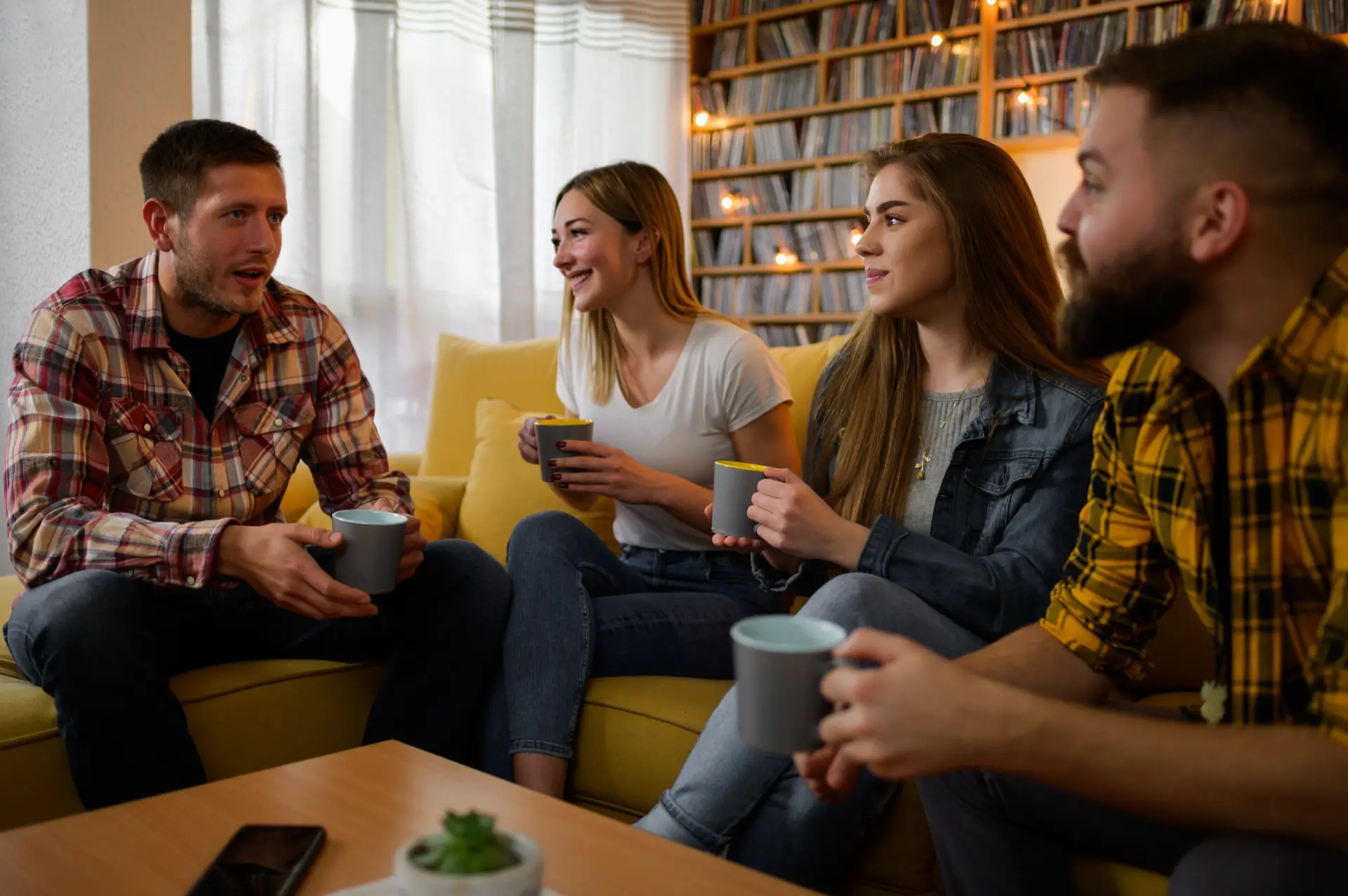Group of friends enjoying a casual conversation on a yellow couch, holding cups, in a cozy living room setting, reflecting support and connection relevant to addiction recovery discussions.