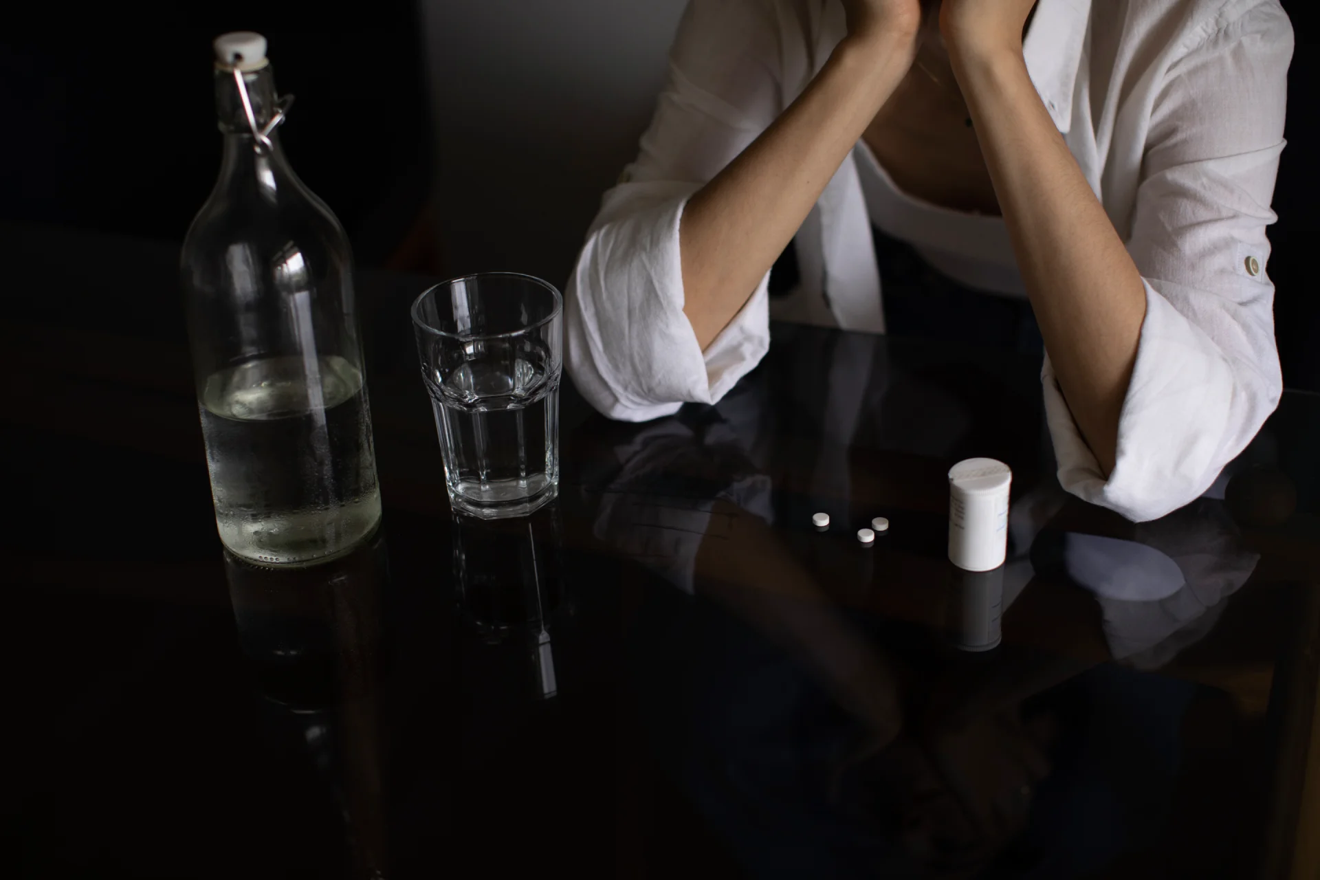 Person with hands on their face, sitting at a table with a glass of water, a bottle of liquid, and pills, illustrating the risks of mixing substances like alcohol and benzodiazepines in the context of GHB addiction.