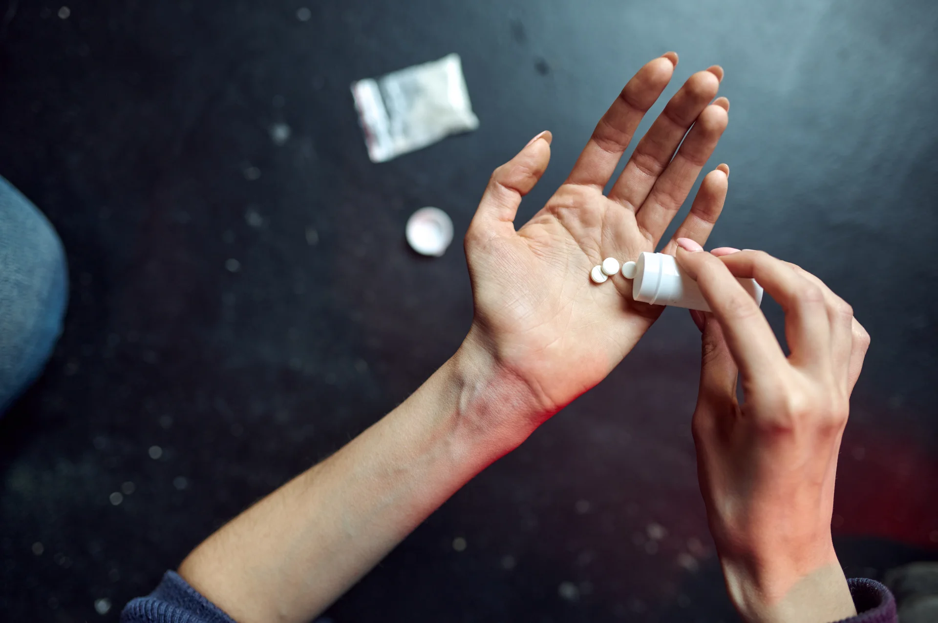 Person pouring white pills from a prescription bottle into their hand, with a small packet visible in the background, illustrating the risks of fentanyl and opioid misuse.