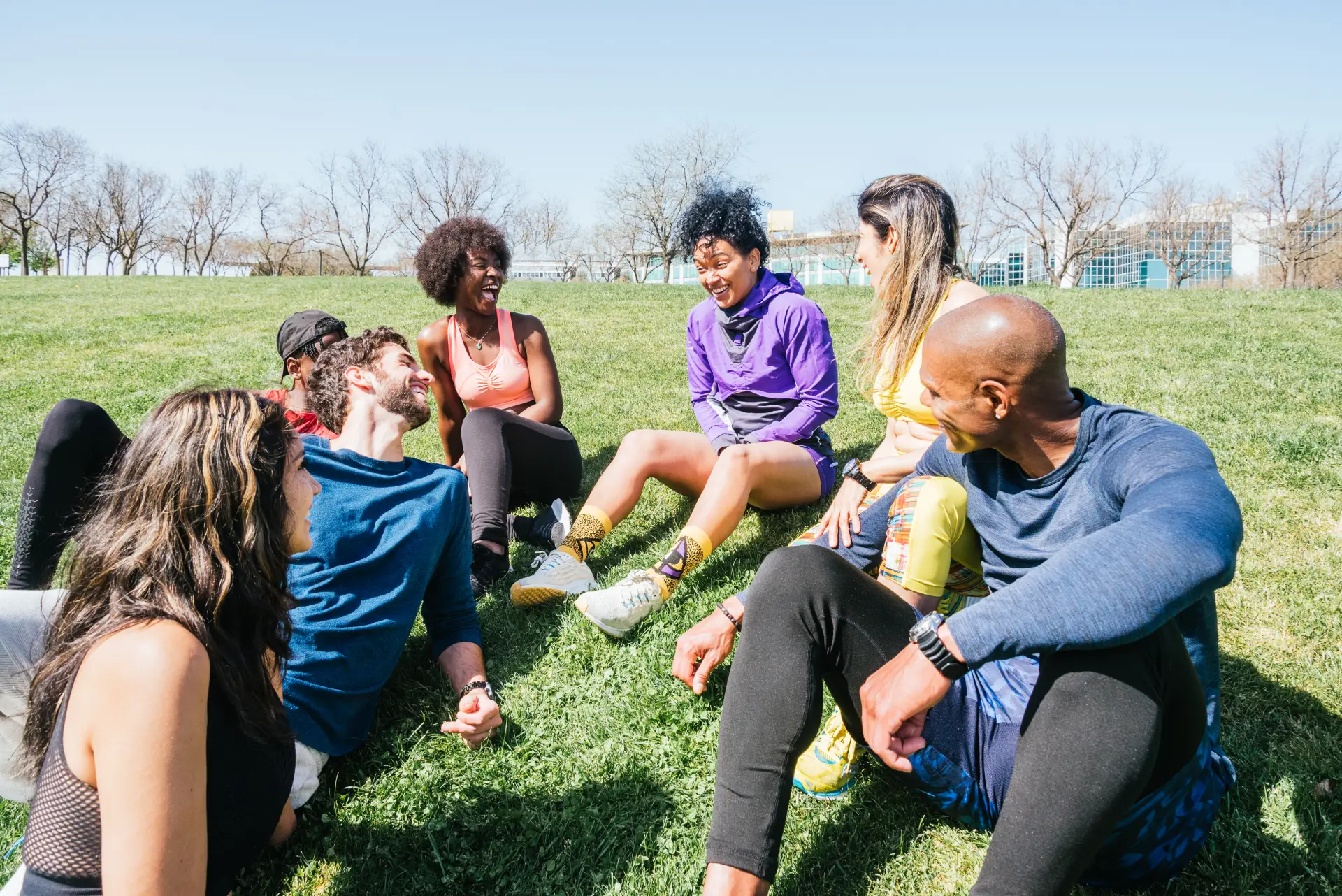 Group of diverse individuals sitting on grass in a park, engaging in conversation and laughter, reflecting community support and connection, relevant to addiction recovery and therapeutic environments.