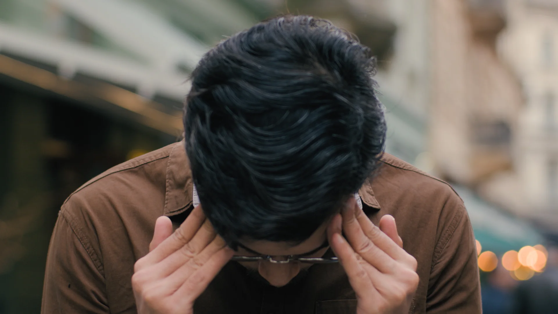 Person with dark hair and glasses holding their head, showing signs of distress, in a blurred outdoor setting, reflecting the emotional impact of PCP addiction discussed in Long Island Addiction Resources.