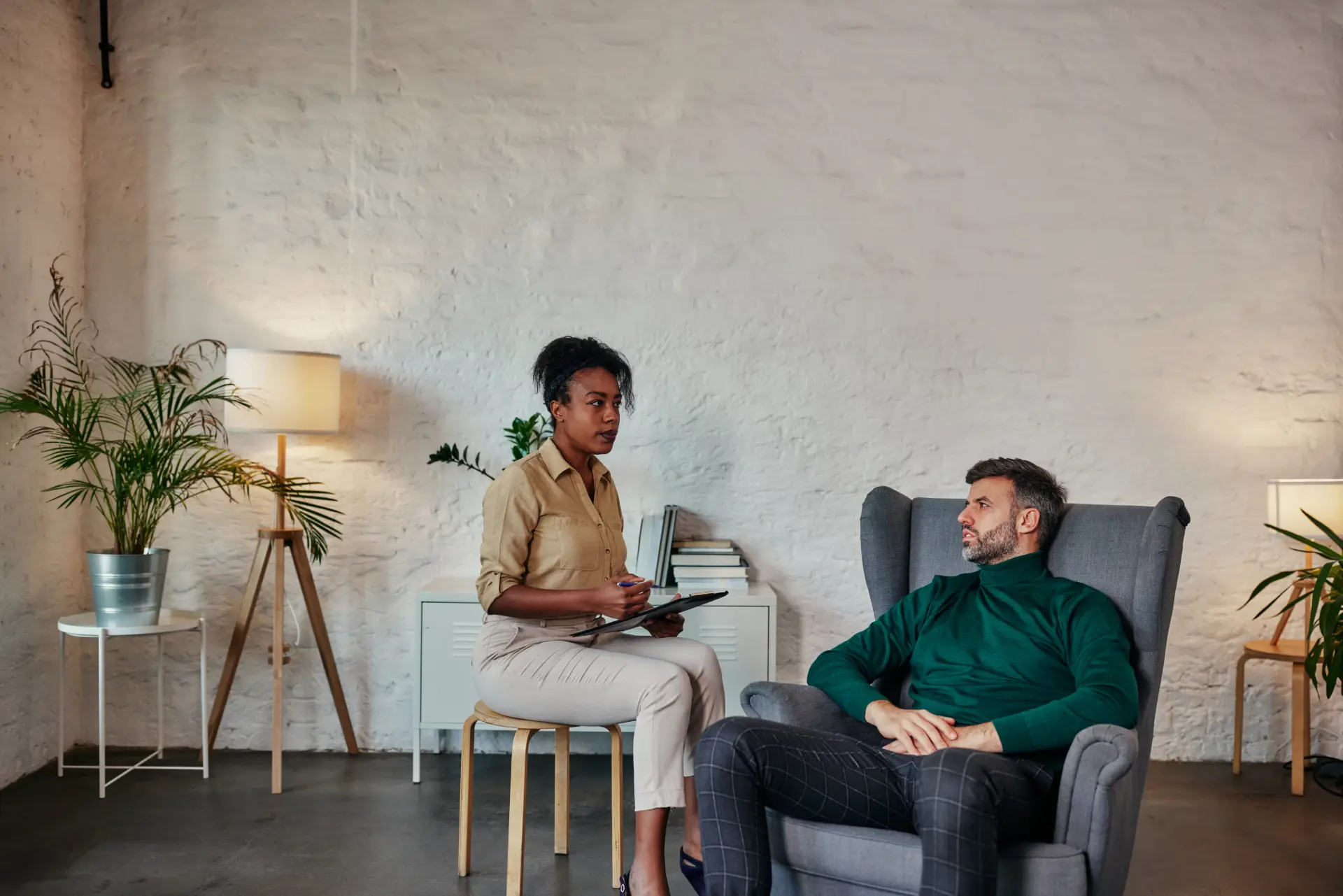 Man sitting in a chair during a therapy session with a female therapist taking notes, discussing alcohol treatment and recovery options in a supportive environment.