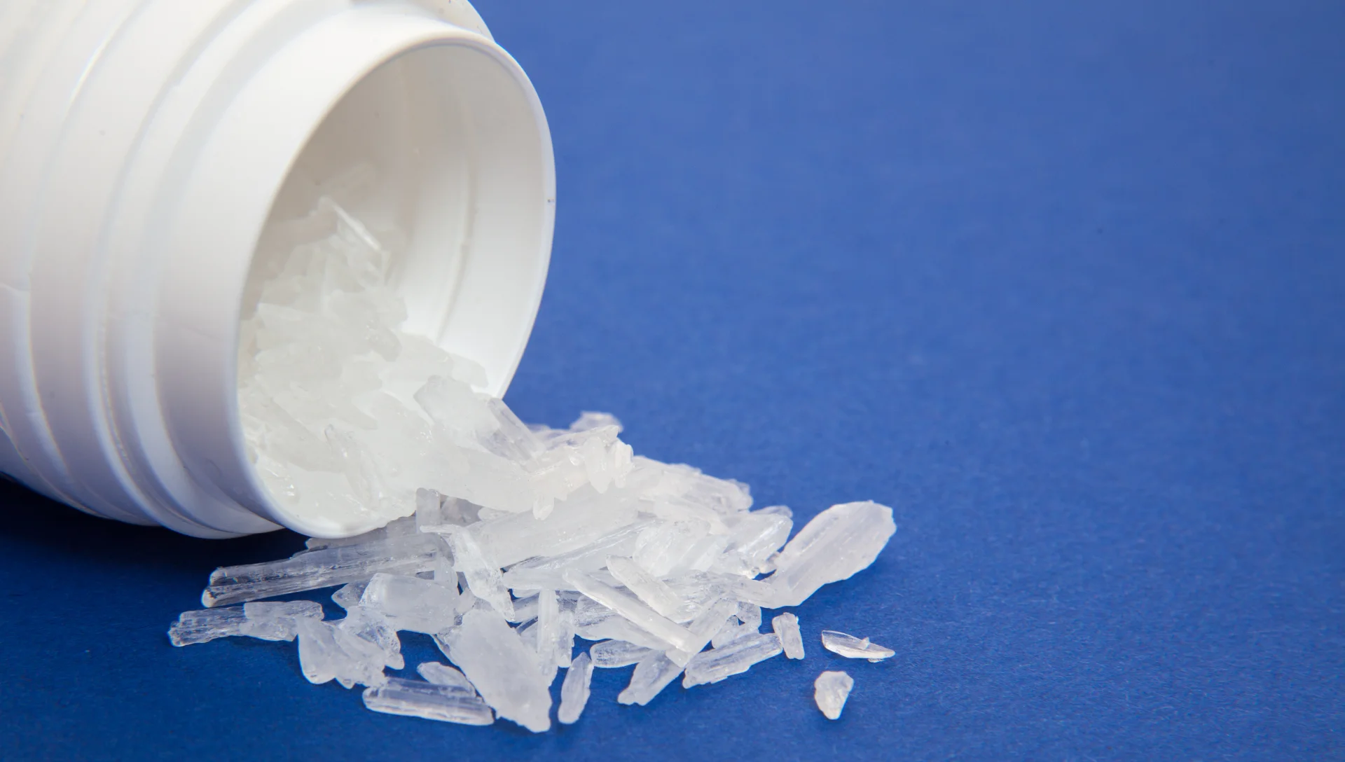 Methamphetamine crystals spilling from a white prescription bottle on a blue background, illustrating the substance's addictive nature and relevance to meth addiction treatment.