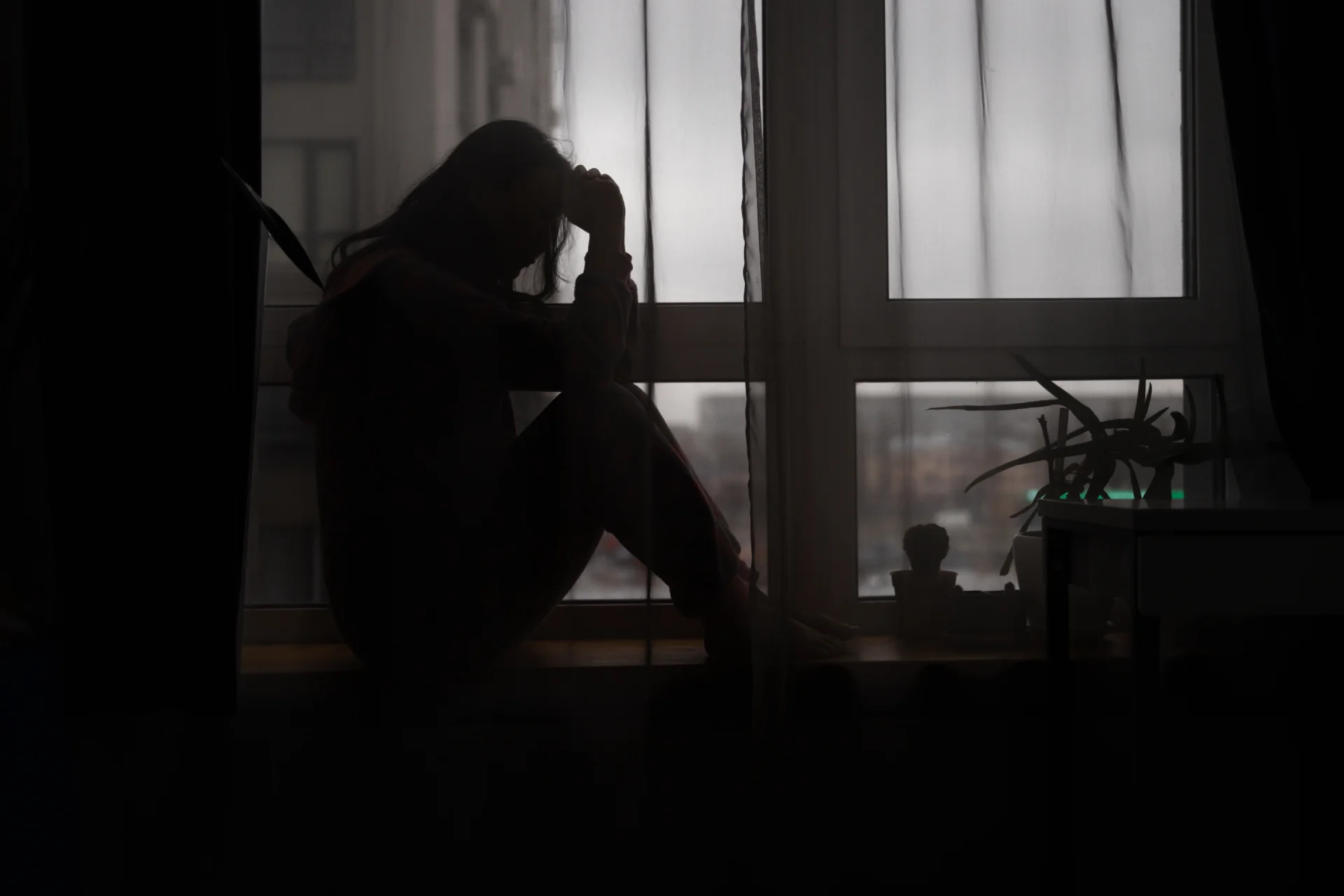 Silhouette of a person sitting by a window, holding their head in distress, surrounded by plants, reflecting themes of depression and suicidal thoughts related to trazodone use.